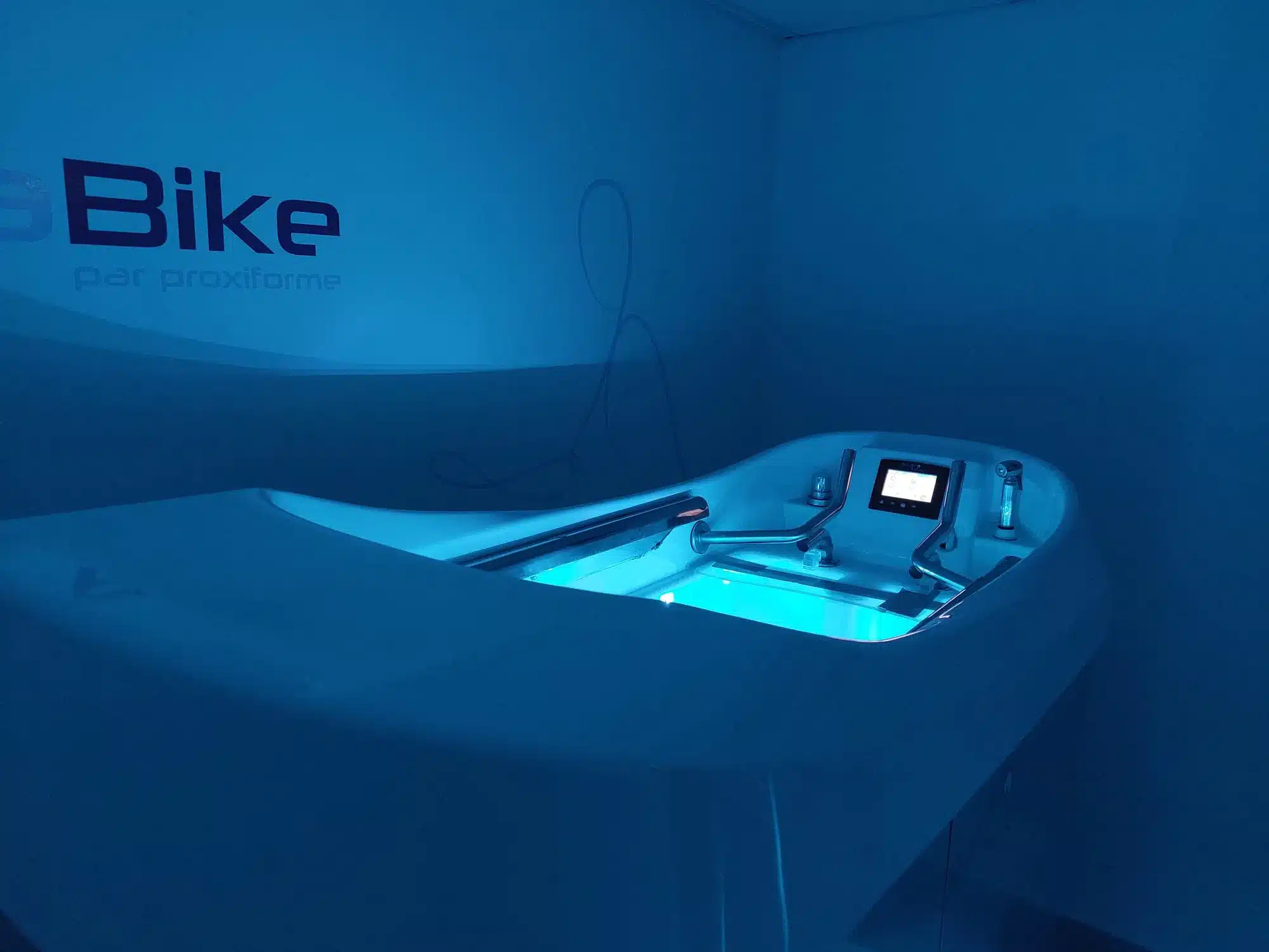 Machine d'hydro-exercice aquatique blanche illuminée en bleu, avec écran et poignées, sous l'inscription 'Bike par proxiforme' sur un mur bleu.