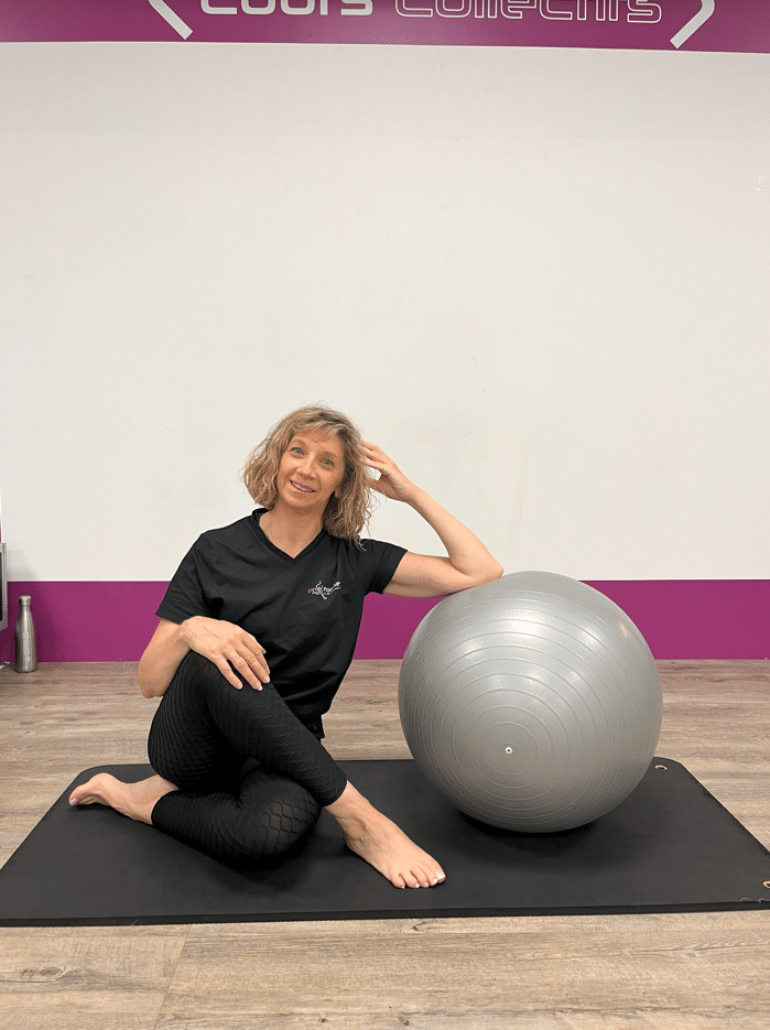 Instructrice de Pilates souriante, vêtue de noir, assise sur un tapis. Elle pose son bras sur un ballon d'exercice gris. Texte : "COURS COLLECTIFS".